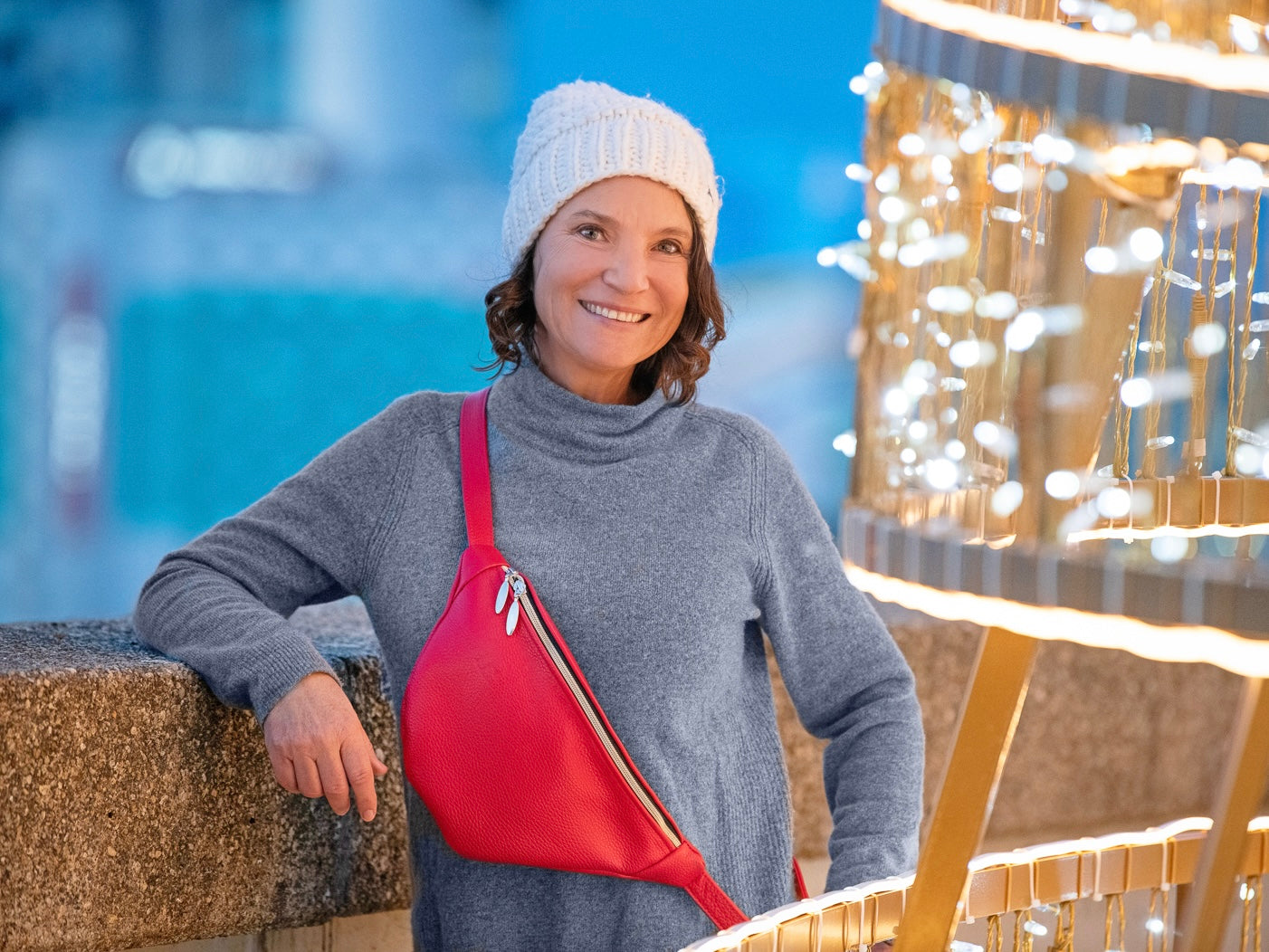 Woman wearing a gray sweater and white beanie with a red bag, standing in front of festive lights.